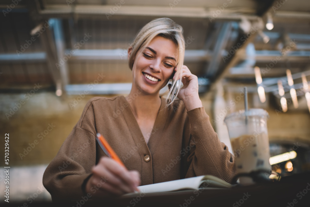 Poster cheerful female student making informative notes while calling via cellphone device for discussing e