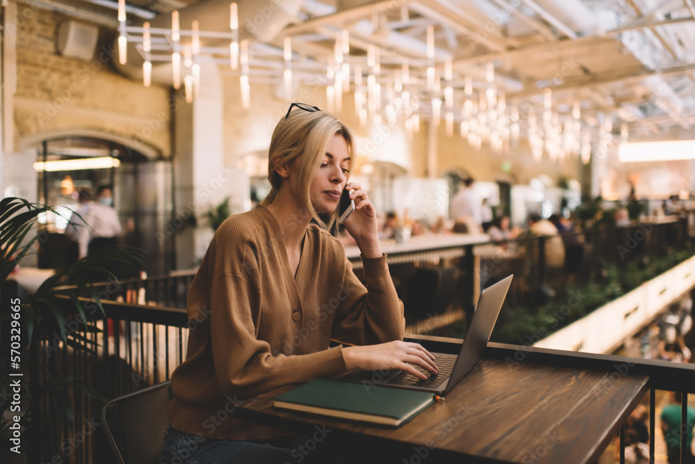 Poster millennial female copywriter using cellphone for call consultancy during laptop browsing in coworkin