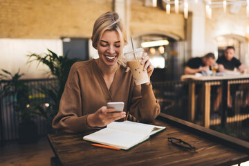 Happy female blogger with ice coffee and textbook planner using mobile technology for online browsing in social networks, cheerful hipster girl connecting to 4g wireless for making booking