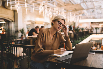 Young female graphic designer in optical eyewear watching webinar video browsed on digital laptop device, millennial hipster girl in spectacles doing distance job while planning content indoors