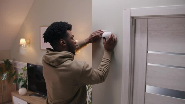African American man puts security camera on wall fastening and connects it to system with cable. Man installs cameras in his apartment. Concept of CCTV cameras, tracking system, safety and privacy.