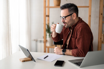 Naklejka premium Freelance worker with glasses in casual clothing holding cup, drinking coffee in home office, sitting near window. Laptop computer, digital tablet device, smart phone, paper document and pen on table.