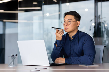 Successful asian man inside office working with laptop, businessman recording audio message using app on smartphone, man using artificial intelligence assistant to help finding a solution.