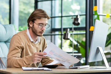 A serious young man accountant, auditor, businessman sits in the office at the table. Concentrated works with documents, calculator, accounts, plans.