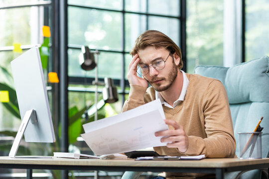 Problems at work. Overtime day. A young pensive and tired businessman sits in the office at the computer, looks at documents, bills, letters in his hands. He holds his head.
