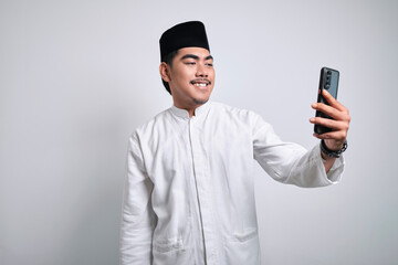 Cheerful young Asian Muslim man doing video call or selfie shot on mobile phone