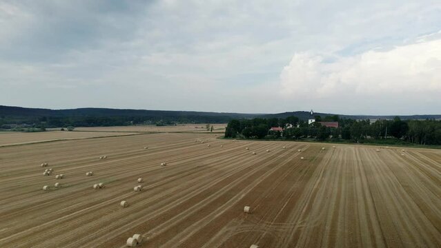 Aerial arc shot of a church surrounded by golden yellow fields with straw bales after harvest. On the background forest and hills on a cloudy sky day. Gammalstorp Kyrka Sweden. Drone high angle view
