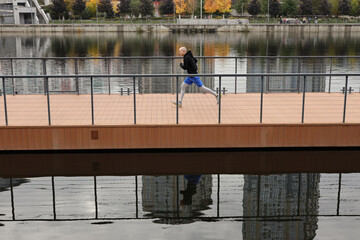 Morning running workout on the bridge near the river. A young bald man in warm sportswear runs in the city embankment. The concept of a healthy lifestyle, fitness, sport.