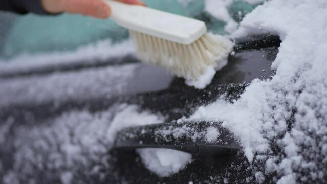 Cleaning snow from car exterior door handle with brush during snowfall cyclone. Snowstorm in winter season. Vehicle care in blizzard