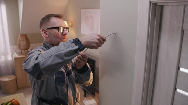 Man in uniform puts cable in drilled hole. Electrician removes insulation from wire with professional tool in apartment. Repair work on electricity. Installation of the Internet or security cameras.