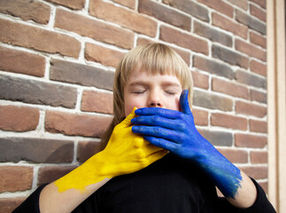 face of child with closed eyes, covered with hands painted in colors of Ukrainian flag