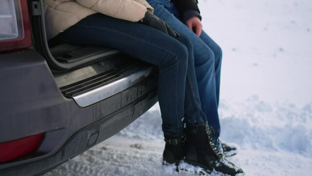 Close up unrecognizable man and woman swaying legs while sitting in open car trunk in snowy field. Family activity on nature in cold season. Traveling journey and healthy active lifestyle concept