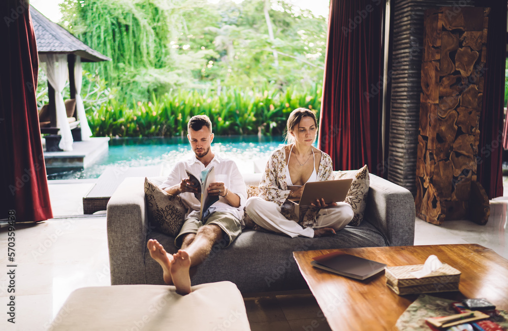 Poster young female freelancer using netbook technology for creating content text while husband reading jou