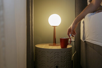Woman in bed reaching for her first morning coffee from the nightstand. A red mug on the bedside...