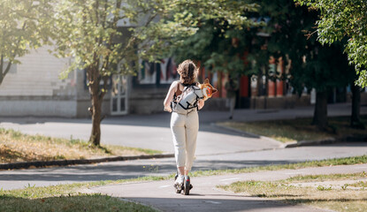 Fototapeta premium Happy smiling woman traveler is riding her electro scooter in city parkland with dog Welsh Corgi Pembroke in a special backpack