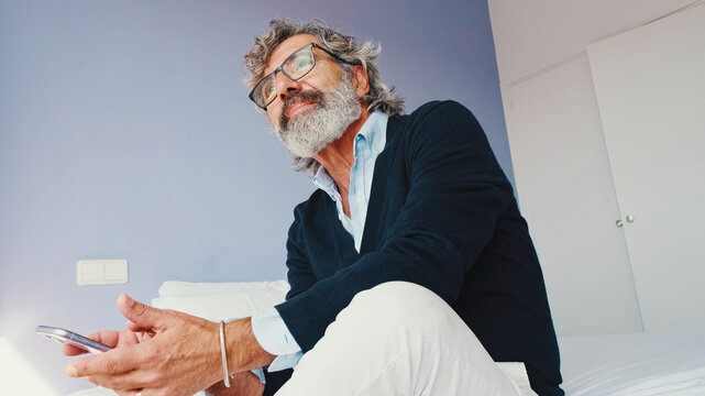 Stylish good looking mature man is sitting on bed and using smartphone