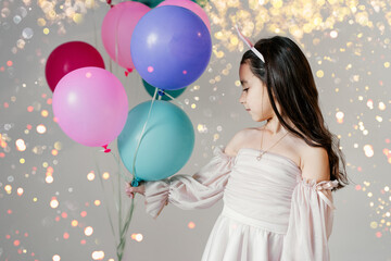 Fototapeta na wymiar Little beautiful shy brunette girl wearing a party hat and pink tutu dress holding balloons in a party. Portrait little pretty child with blue, green and pink balloons.