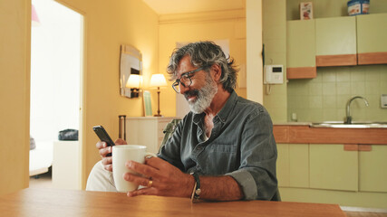 Elderly man drinks coffee from mug and uses mobile phone at home