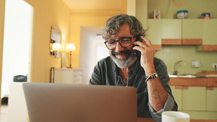 Mature businessman working on laptop, talking on mobile phone sitting in home kitchen
