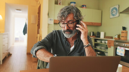 Mature businessman working on laptop, talking on mobile phone sitting in home kitchen