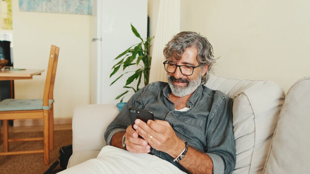 Middle-aged man relaxing on his couch while in home and using his smartphone to text