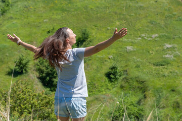 Happy woman greeting sun in ravine covered with greenery. Young female raise hands to the sunshine. She looking on valley and showing  freedom gesture. Positive joyful of emotion surya namaskar.