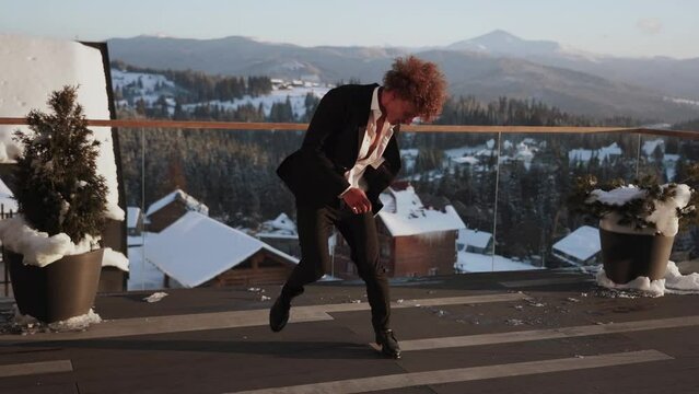 A stylish young male dancer in a classic black suit dances against the backdrop of snowy mountains on the terrace.The concept of modern dance