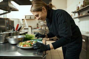 Smiling female chef in restaurant kitchen decorates plate with salad