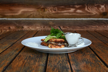 Pancakes from grated potatoes with mushrooms and sour cream on a white plate on a wooden background.