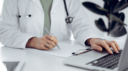 Fototapeta premium Unknown doctor woman sitting and writing notes at the desk in clinic or hospital office, close up. Medicine concept