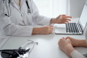 Doctor and patient sitting at the desk in clinic office. The focus is on female physician's hands pointing to laptop computer, close up. Medicine and helthcare concept - 570408600