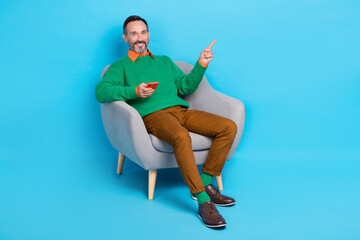 Fototapeta na wymiar Full size photo of mature attractive male gadget point empty space armchair dressed stylish green outfit isolated on blue color background