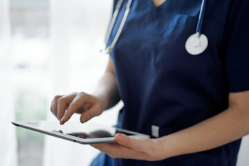Doctor woman using tablet computer while standing near panorama window in clinic, close up. Physician or surgeon at work. Medicine concept