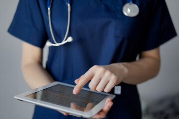 Doctor woman using tablet computer while standing near panorama window in clinic, close up. Physician or surgeon at work. Medicine concept