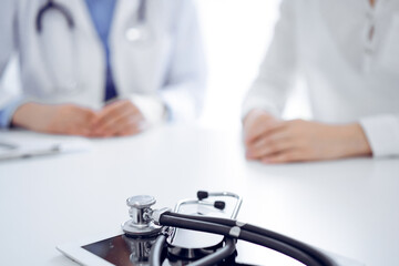 Stethoscope lying on the tablet computer in front of a doctor and patient sitting near of each other at the background. Medicine, healthcare concept