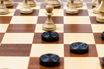 playing by different rules on the same board - black checkers and white chess figures on wooden chessboard, pawn and checkers piece in center of playfield closeup (focus on the pawn)