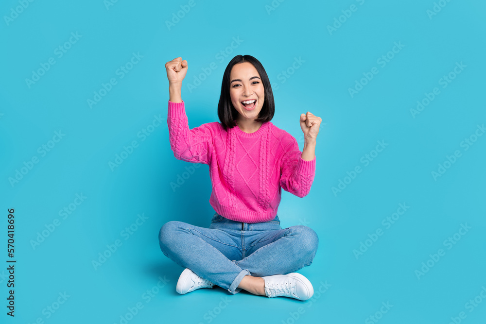 Wall mural Full length photo of lucky shiny woman dressed pink sweater rising fists sitting legs crossed isolated blue color background