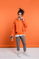 Fototapeta premium pierced and multiracial man with wireless headphones holding paper cup with coffee to go on coral background