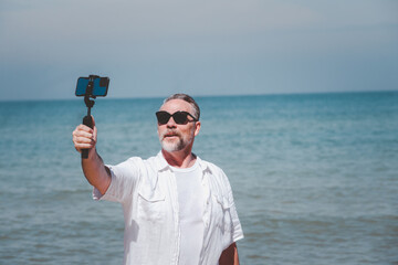 elderly men travel and relax at the beach on a clear day. He uses his smartphone to film himself. Make a vlog or maybe a video call to his family
