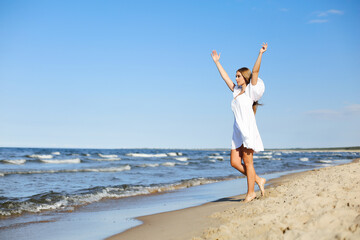 Happy, beautiful woman on the ocean beach standing in a white summer dress, raising hands - Powered by Adobe