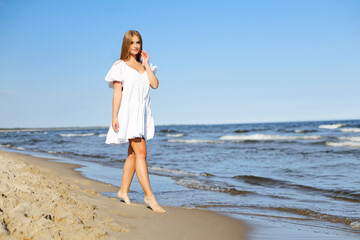 Happy smiling beautiful woman is walking on the ocean beach in a white summer dress