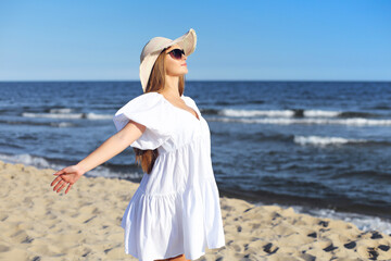 Fototapeta na wymiar Happy blonde woman is on the ocean beach in a white dress and sunglasses, open arms