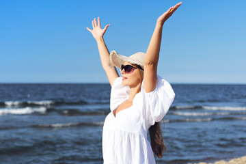 Fototapeta na wymiar Happy blonde woman is on the ocean beach in a white dress, sunglasses and hat, raising hands