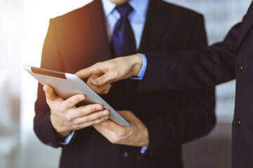 Business people use a tablet computer for discussion of their new project, standing in sunny modern office. Unknown businessman or male entrepreneur with a colleague at workplace