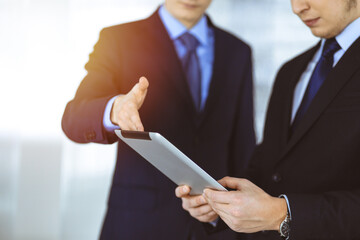 Business people use a tablet computer for discussion of their new project, standing in a sunny modern office. Unknown businessman or male entrepreneur with a colleague at workplace