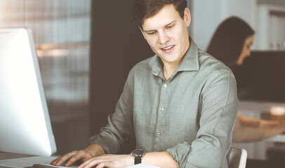 Young businessman working on his computer sitting at the desk in office. Headshot portrait of a man