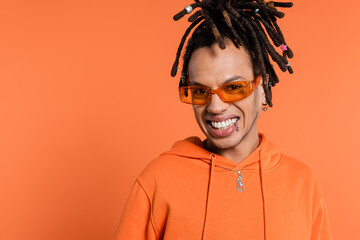 happy multiracial man with dreadlocks wearing stylish sunglasses isolated on coral background