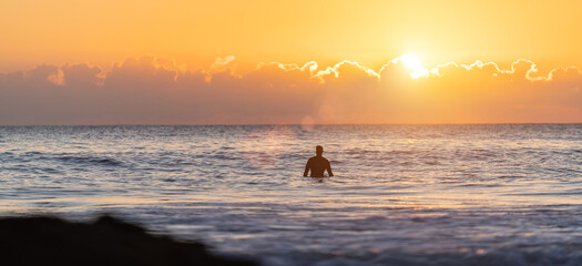 surfer sitting on their surfboard in the blue ocean and waiting for a wave at sunset in the evening...