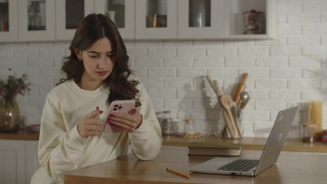 A brunette woman sitting in her home office uses her phone to search for information that will help her solve a work task. Remote work using phone and laptop. Girl scrolling on smartphone screen