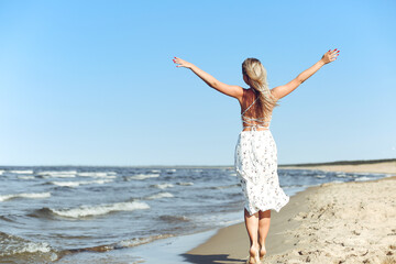 Happy blonde beautiful woman on the ocean beach standing in a white summer dress, open arms
